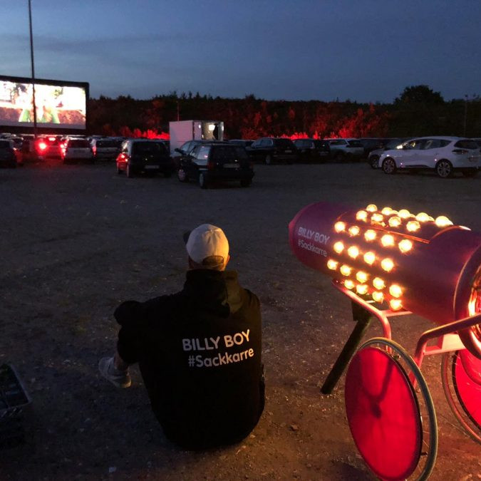 Billy Boy Autokino: Der Promoter sitz neben der beleuchteten Sackkarre auf dem Boden und guckt ein Film im Autokino.