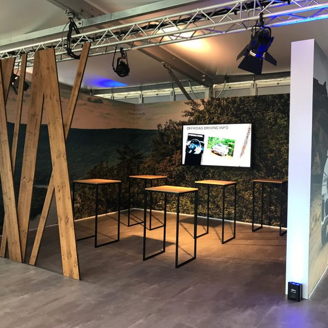 BMW Group: In einem abgetrennten Teil des Zeltes stehen Stehtische mit einer Holzplatte. An der Wand hängt ein TV auf dem man Offroad Driving Infos bekommt.