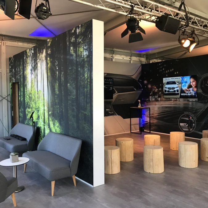 BMW Group: auf der rechten Seite ist eine Tagunssituation, in der die Gäste auf abgesägten Baumstämmen sitzen und links ist eine Lounge, in der man auf grauen Sofas sitzt.