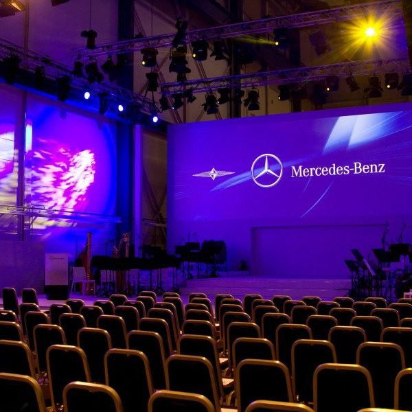 Daimler 75 Jahre PKW: Eine leicht beleuchtete Orchesterbühne mit leeren Stuhlreihen davor.