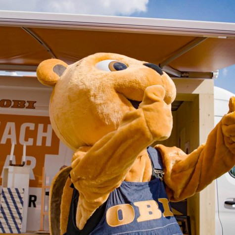 OBI MachBar Roadshow: Das OBI Maskottchen. Es Steht mit beiden Daumen nach oben vor einem OBI Sprinter.