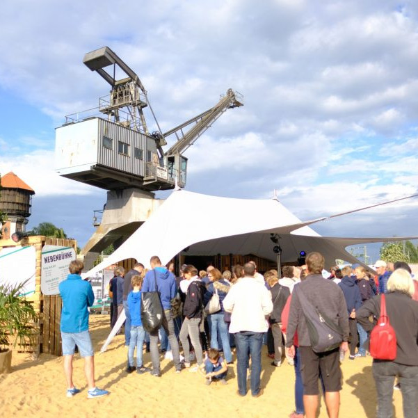 OLB 150 Jahre Festival: Geladene Gäste stehen im Sand vor einem Zelt, im Hintergrund ist ein alter Kran.