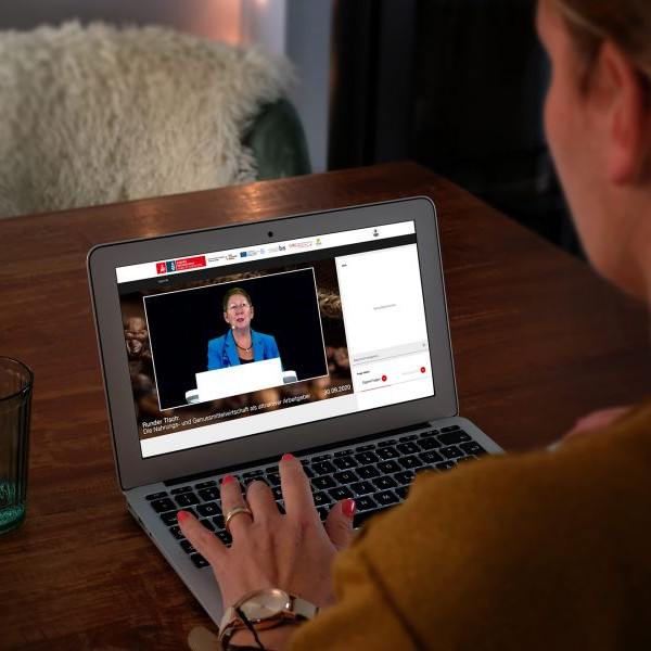Nahrungs- und Genussmittelwirtschaft: Blick über die Schulter einer Onlineteilnehmerin die vor ihrem Laptop sitzt. Im Livestream ist eine Rednerin zu sehen.